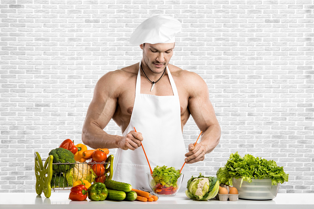 Een gespierde man bereid in niet veel meer kleding dan een schort en koksmuts, bereidt een salade.